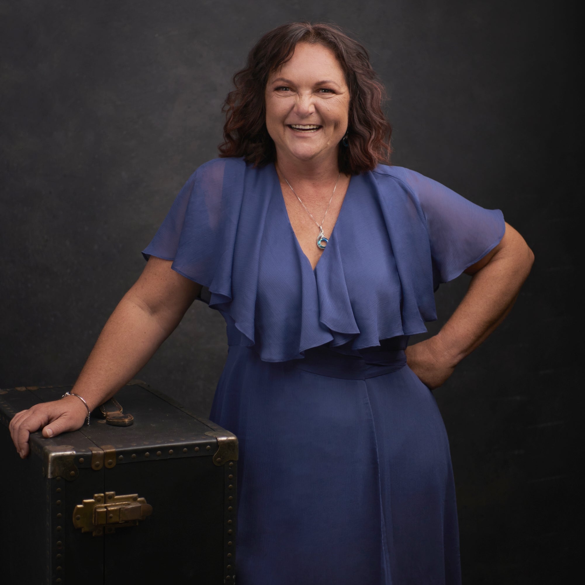 A smiling woman with shoulder-length brown hair stands in a studio wearing a blue ruffled short-sleeve dress and leaning on a dark vintage trunk against a dark gray backdrop.