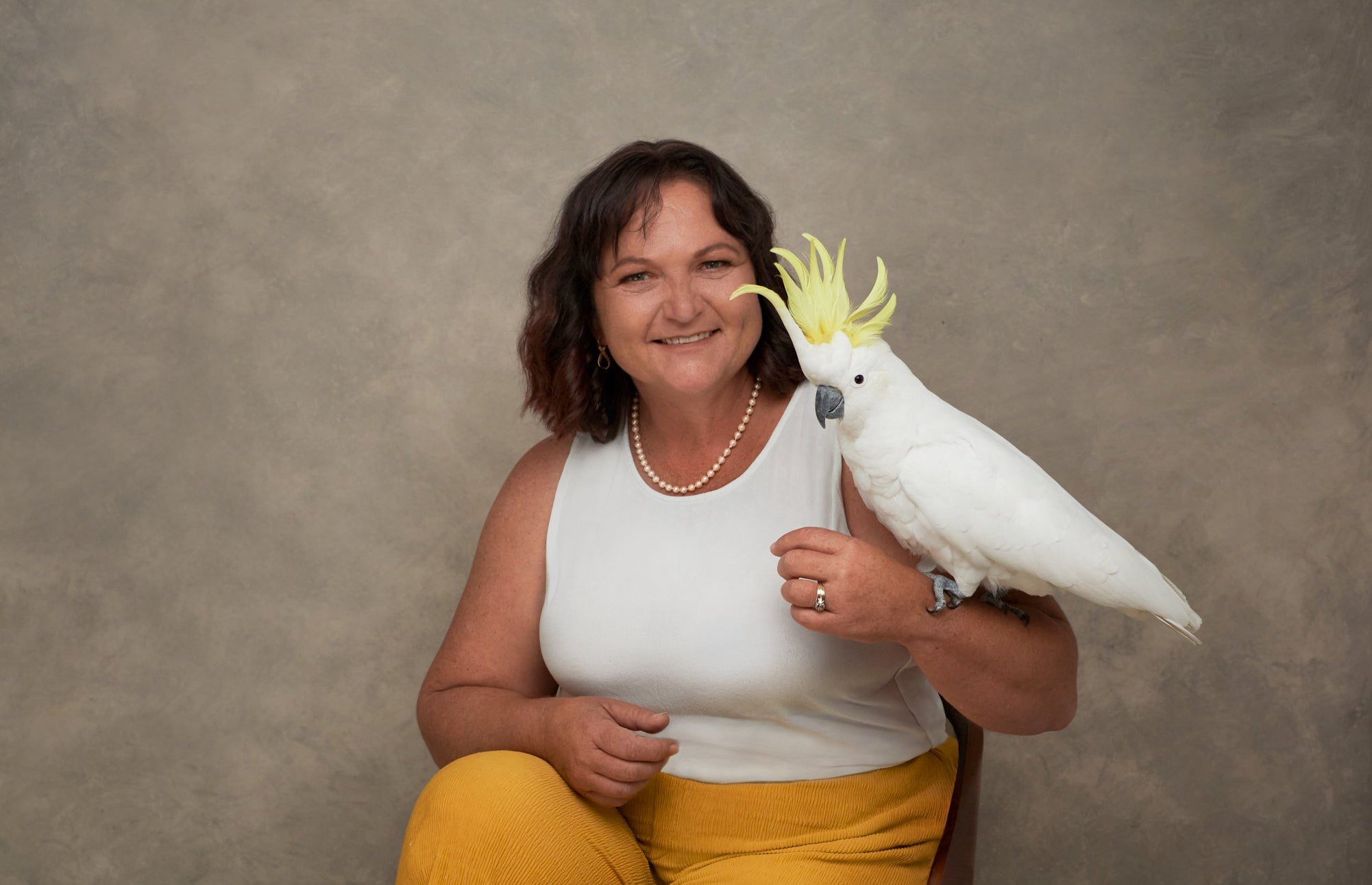 A smiling woman with dark hair sits against a textured gray studio backdrop, holding a white cockatoo with a bright yellow crest. She wears a white sleeveless top and mustard yellow pants.