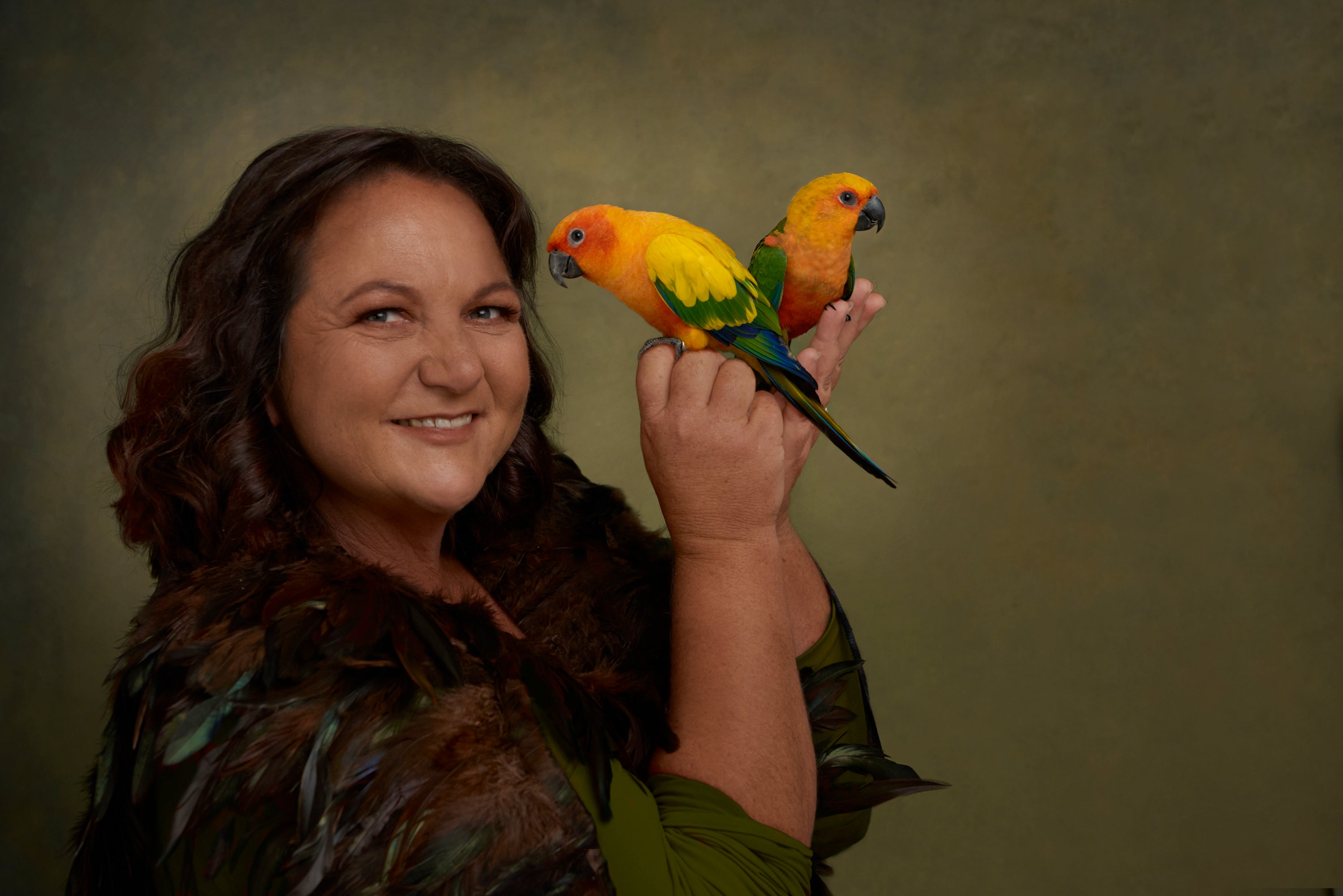 A smiling woman with long dark hair holds two bright conure parrots on her hands against a textured olive-green studio background.