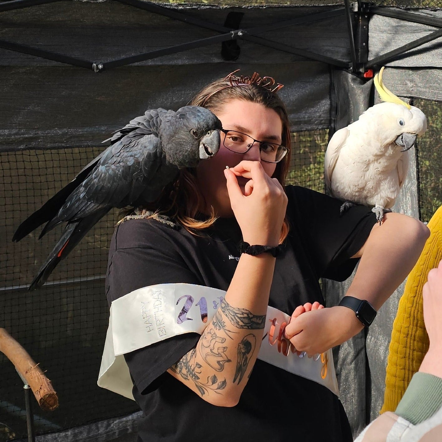 A woman wearing glasses and a black T-shirt with a white sash has two parrots, one black and one white, on her shoulders in an outdoor event setting under a canopy.